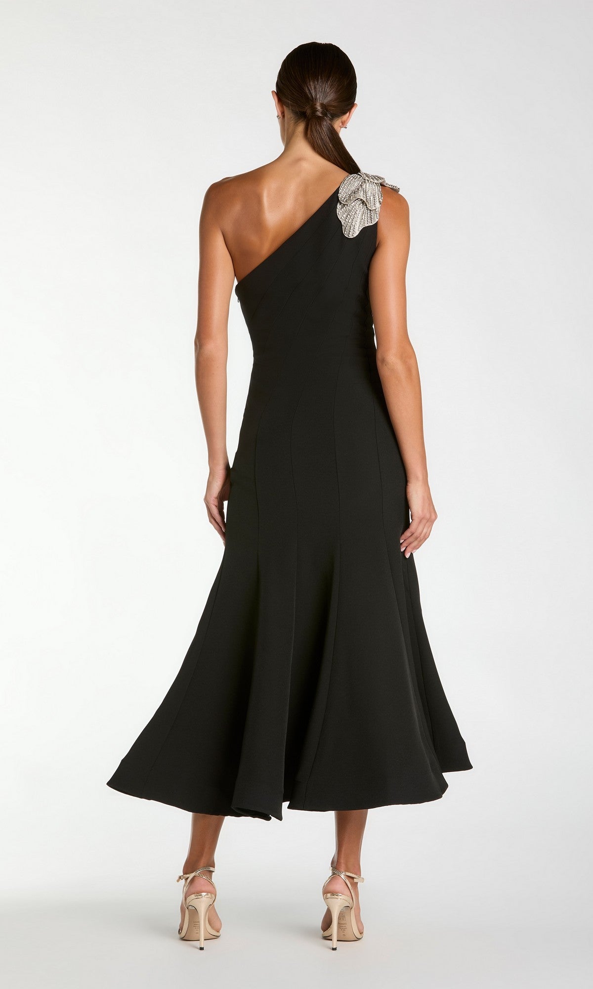 A woman stands with her back to the camera, wearing the Tea-Length Party Dress: Mac Duggal 13221 in black crepe, featuring a one-shoulder design adorned with an embellished floral shoulder applique. She pairs the dress with beige high-heeled sandals.